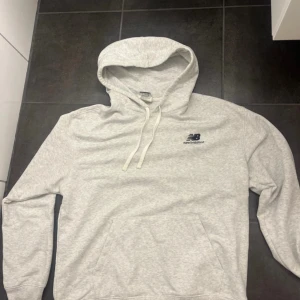 Ljusgrå hoodie från New Balance - Snygg ljusgrå hoodie från New Balance med broderad logga på bröstet. Tröjan har huva med snörning, känguruficka och långa ärmar. Perfekt för chill eller träning, riktigt skön och stilren. Materialet är mjuk bomullsmix.
