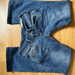 Tommy Hilfiger blå bootcut jeans - Snygga blå jeans från Tommy Hilfiger med bootcut-modell och låg midja. Klassisk femficksdesign, dubbla knappar i midjan och detaljerade bakfickor med Hilfiger-logga.