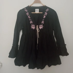 Svart klänning med broderi Love Forever - Svart kortklänning från Love Forever med långa ärmar, v-ringning och rosa broderade blommor vid halsen. Klänningen har snörning framtill och volangdetaljer vid ärmslut och nederkant. Perfekt för dig som gillar boho-stil och unika detaljer.