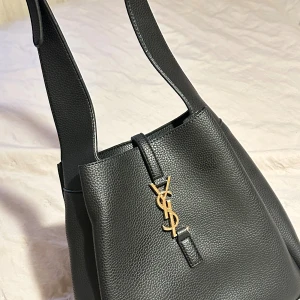 Svart axelväska från Saint Laurent - Säljer en svart axelväska från Saint Laurent i exklusivt skinn med guldigt YSL-emblem framtill. Väskan har en stilren, strukturerad form, bred axelrem och innerfack med dragkedja. Perfekt för dig som vill ha en lyxig och tidlös accessoar.