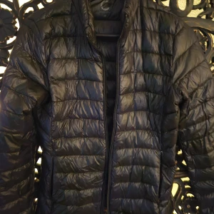 Svart pufferjacka med dragkedja Cubus - Säljer en svart pufferjacka med quiltad design och hel dragkedja framtill. Jackan har lång ärm och en clean, sportig look som passar perfekt när det är kallt ute. Materialet är glansigt och lätt, vilket gör den smidig att bära.