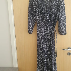 Mörkblå blommig omlottklänning - Säljer en lång, marinblå omlottklänning med vitt blommönster. Klänningen har långa ärmar och volangdetalj vid ringningen. Den har använts 2 gånger sedan den köptes