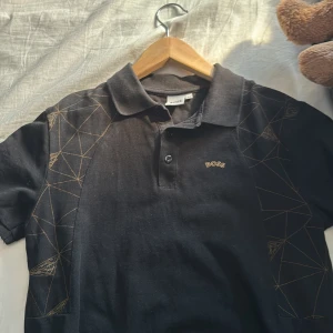 Svart BOSS pikétröja med mönster - Svart pikétröja från BOSS med coola geometriska linjemönster i guld på axlar och ärmar. Klassisk krage, knäppning framtill och diskret BOSS-logga på bröstet. Perfekt för dig som vill ha en stilren men unik look.