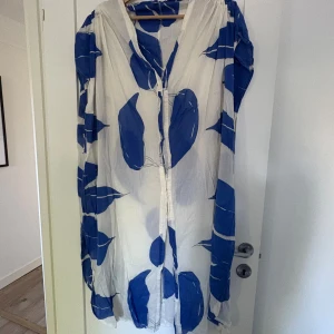 Vit och blå strandklänning med mönster - Strandklänning i tunn bomull med vitt tyg och stora blå bladliknande mönster. Klänningen har detaljer vid axlarna och knäppning framtill. Luftig och flowy passform, perfekt för varma dagar.