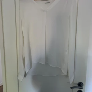 Vit blus med v-ringning från VILA - Säljer en stilren vit blus från VILA med v-ringning och långa ärmar. Blusen har en lätt och flowy känsla, knappar vid ärmsluten och en något längre bakdel. Perfekt att styla med jeans eller kjol för en clean look.