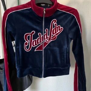 Marinblå och vinröd velourtröja Jaded Ldn - Säljer en snygg marinblå och vinröd velourtröja från Jaded Ldn med dragkedja framtill och broderad logga. Tröjan har långa ärmar med röda ränder och vit piping längs axlarna. Perfekt för dig som gillar sportig och trendig stil.Storleken passar XS/S