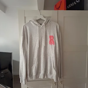 Grå hoodie från Risky med rosa patch - Grå hoodie från Risky med stor rosa 'R'-patch på bröstet och tryckt Risky-logga på huvan. Tröjan har känguruficka, dragsko i huvan och en relaxed passform. Perfekt för dig som gillar streetwear och snygga detaljer.