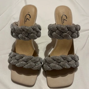 Glittriga sandaler med flätade band - Snygga sandaler från Clou i beige med två breda, flätade band täckta av silverfärgat strass. Skorna har fyrkantig tå och en låg klack. Storlek 39, endast provade.