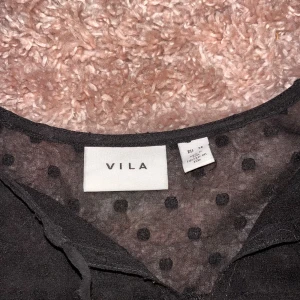 Svart prickig blus från Vila - Säljer en svart blus från Vila med transparenta partier och prickigt mönster. Blusen har långa ärmar med lätt volym och fina spetsdetaljer vid axlar och ärmslut. Perfekt för dig som gillar en stilren men ändå lekfull look.