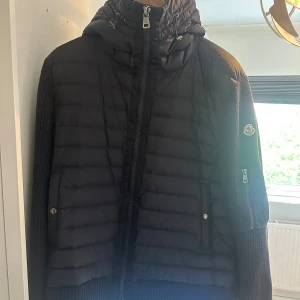 Mörk blå dunjacka från Moncler - Mörk blå dunjacka från Moncler med stickade ärmar och vadderad kropp. Jackan har huva med dragsko, dragkedja framtill och Moncler-logga på ärmen. Perfekt för kyliga dagar och snygg till både jeans och sneakers. Priset är inte hugget i sten 