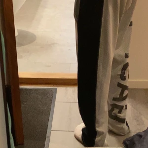 Svarta och vita mjukisbyxor med tryck - Säljer ett par mjukisbyxor i svart och vitt med breda kontrastpaneler och tryckt text längs ena benet. Byxorna har avslappnad passform och är perfekta för chill dagar hemma eller streetwear-stil. Materialet är mjukt och andas.