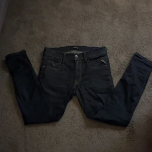 Replay Anbass mörkblå jeans W31 L30 - Snygga Replay Anbass jeans i mörkblå tvätt med klassisk femficksdesign och smal passform. Jeansen har dragkedjegylf, läderpatch bak och diskreta kontrastsömmar. Perfekta för dig som gillar stilrena och moderna jeans.