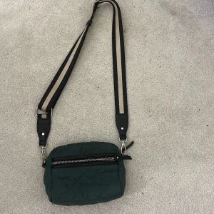 Grön quiltad axelväska med randigt band - Snygg mörkgrön quiltad axelväska med bredt randigt axelband i svart och beige. Väskan har en dragkedja framtill och ett coolt leopardmönstrat innerfoder med flera fack. Perfekt storlek för mobilen och småsaker, och ger en trendig touch till din outfit.