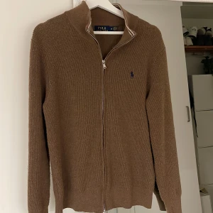 Brun stickad kofta från Polo Ralph Lauren - Snygg brun stickad kofta från Polo Ralph Lauren med dragkedja framtill och klassisk broderad logga på bröstet. Koftan har ribbade muddar och hög krage, perfekt för lager-på-lager. Passar dig som gillar stilren och tidlös design.