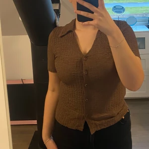 Brun kortärmad blus med knappar - Säljer en brun tröja med korta ärmar och v-ringning. Toppen har en krage och knappar hela vägen framtill. Perfekt att styla med jeans eller kjol. Helt ny