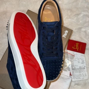 Louboutin skor - Säljer ett par mörkblå sneakers från Christian Louboutin i mocka med ikoniska röda sulor. Skorna har snörning och coola nitar framtill på tån. Kommer med originalkartong och dustbag. Perfekta för dig som vill sticka ut med lyxig streetstyle.