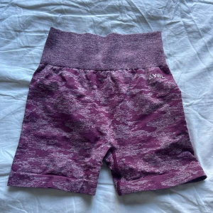 Lila camo träningsshorts från AYBL - Säljer ett par lila camo-mönstrade shorts från AYBL med hög midja och tight passform. De är tillverkade i ett stretchigt syntetmaterial som passar perfekt för träning. Shortsen har en bred, ribbad midjeresår och diskret logga framtill. Även scrunch baktill! 