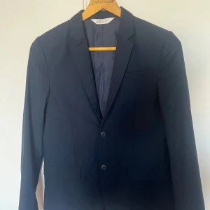 Mörkblå kavaj från H&M, storlek 170 - Säljer en stilren mörkblå kavaj från H&M i storlek 170. Kavajen har två knappar framtill, klassisk krage och två fickor med lock. Perfekt för dig som vill ha en enkel och snygg look till speciella tillfällen.