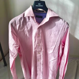 Ljusrosa skjorta från GANT - Säljer en klassisk ljusrosa skjorta från GANT med knappar framtill och krage. Skjortan är långärmad och tillverkad i bomull, perfekt för en clean och stilren look. Passar dig som gillar tidlös stil och vill ha något som sticker ut lite extra.