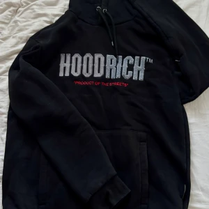 Svart HOODRICH hoodie - Svart hoodie från HOODRICH i storlek M med stor broderad logga i grått och vitt samt röd text på bröstet. Klassisk känguruficka, snörning i huvan och mjukt material. I bra skick, inga skador.