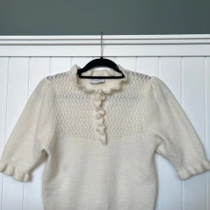 Vit stickad blus från Holly & Whyte - Supersöt vit stickad blus från Holly & Whyte med volangkant vid halsen, pärlknappar och korta puffärmar. Överdelen har ett diskret hålmönster och blusen är i mjukt material, perfekt för lager-på-lager eller som statement-piece.