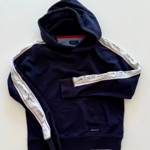 Marinblå hoodie från Gant - Snygg marinblå hoodie från Gant med vita ränder och logga längs ärmarna. Tröjan har huva, magficka och ribbade muddar. Perfekt för en avslappnad och sportig stil.