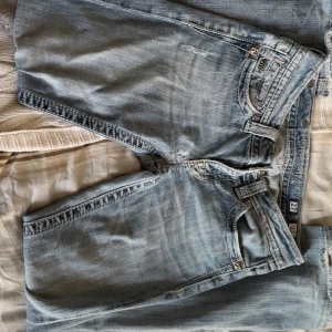 Miss Me ljusblå jeans med dekorerade fickor - Ljusblå jeans från Miss Me med snygga slitningar och broderade bakfickor med svarta och silvriga detaljer. Klassisk femficksmodell med låg midja och utvängda ben. Jeansen har coola detaljer och är perfekta för dig som gillar 2000s stil