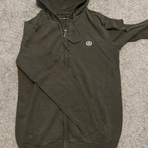 Mörkgrön hoodie från Henri Lloyd - Snygg mörkgrön hoodie från Henri Lloyd med dragkedja och huva. Tröjan har broderad logga på bröstet, ribbade muddar och praktiska fickor framtill.