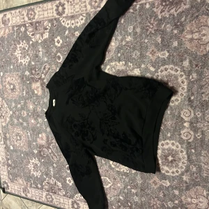 Svart sweatshirt med blommigt mönster - Svart sweatshirt från YOURTURN med diskret sammetsliknande blommigt mönster över hela tröjan. Klassisk rund halsringning och långa ärmar. Perfekt för dig som gillar stilrena men ändå unika detaljer.