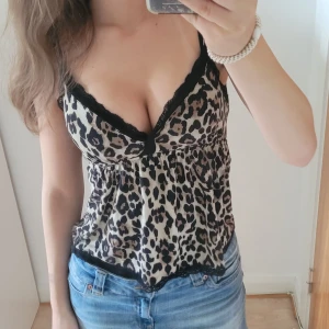 Leopardmönstrad topp med spets - Säljer en snygg leopardmönstrad linnetopp med svarta spetsdetaljer vid halsringning och nederkant. Toppen har smala axelband och en v-ringad form som ger en feminin look. Materialet är mjukt och lätt, perfekt för att sticka ut med ett coolt mönster.