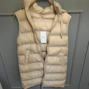 Lång beige dunväst  - En helt ny beige dunväst från Vila. Storlek 34. Perfekt nu till hösten