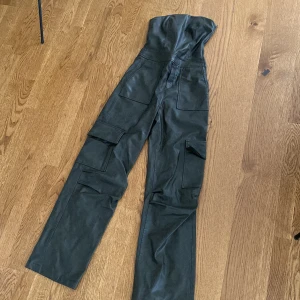 Svart byxdress i skinnimitation Zara XS - Supersnygg svart byxdress från Zara i skinnimitation. Högmidjad modell med raka ben, flera stora cargofickor och dragkedja i sidan. Perfekt för dig som vill sticka ut med en edgy och trendig look.
