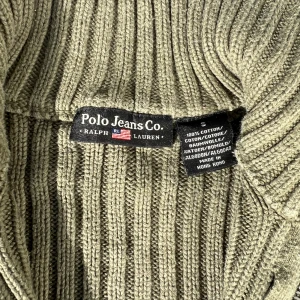 Olivgrön ribbad kofta från Polo Jeans - Säljer en olivgrön ribbstickad kofta från Polo Jeans Co. med hel dragkedja och hög krage. Tröjan är i 100% bomull och har lång ärm. Perfekt för lager-på-lager och chill stil.