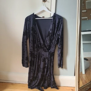 Svart blommig omlottklänning Lindex - Supersnygg svart omlottklänning från Lindex med blommigt mönster. Klänningen har lång ärm, v-ringning och knyt i midjan. Mjuk och stretchig känsla, perfekt för dig som gillar en feminin och bekväm stil.