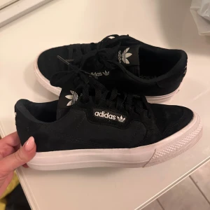 Svarta Adidas sneakers med vit sula - Snygga svarta Adidas sneakers med vit sula och klassisk logga på sidan och plösen. Skorna har snörning och är tillverkade i mocka och textil för en stilren look. Perfekta för dig som gillar streetstyle och vill ha något som funkar till det mesta.