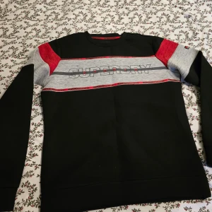 Superdry svart och röd sweatshirt - Snygg svart sweatshirt från Superdry med breda grå och röda paneler över bröst och ärmar. Logotypen är tryckt framtill och röda detaljer ger en sportig vibe. Tröjan har långärmad passform och är gjord i mjuk bomullsmix. Perfekt för dig som gillar streetwear.