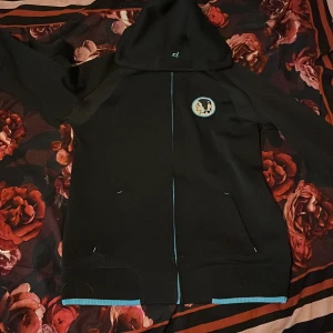 Svart hoodie Peak Performance S - Svart hoodie från Peak Performance med ljusblå detaljer och dragkedja. Har huva, två fickor och broderad logga på bröstet. Snyggt tryck på ena ärmen och ribbade muddar med blå kant. 