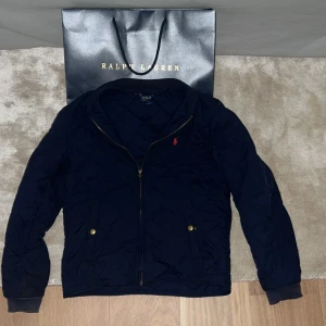 Mörkblå quiltad jacka Polo Ralph Lauren - Quiltad mörkblå jacka från Polo Ralph Lauren med guldfärgade knappar på fickorna och dragkedja framtill.  Perfekt för kyliga höst dagar och snygg till jeans. Priset är ej hugget i sten!