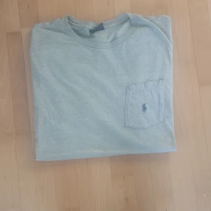 Ljusgrå t-shirt från Polo Ralph Lauren - Snygg ljusgrå t-shirt från Polo Ralph Lauren med klassisk passform. T-shirten har en bröstficka med broderad blå logga och är tillverkad i 100% bomull. Perfekt basic-plagg med enkel rund hals och korta ärmar. Priset är såklart inte hugget I sten😉😉