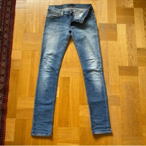 Nudie Jeans Slim Blå 28/32 - Säljer ett par blå slim jeans från Jeansen har klassisk femficksdesign, orangea sömmar och Nudie-logga på bakfickan. De är tillverkade i stretchig bomullsdenim för en lösare passform och har snygg tvätt på låren. 