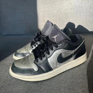 Nike Air Jordan 1 Low svart/silver - Säljer ett par Nike Air Jordan 1 Low i svart och silver. Skorna har snörning, rund tå och platt sula. Ovandelen är i skinn och textil, perfekt för dig som gillar streetwear. Väldigt fräscha