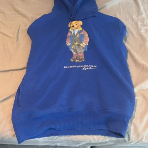 Blå Polo Bear hoodie från Ralph Lauren - Blå hoodie från Polo Ralph Lauren med ikoniskt Polo Bear-tryck på bröstet. Tröjan har huva med snörning och känguruficka. Mjuk bomullskänsla och klassisk passform, perfekt för dig som gillar streetwear och vill sticka ut med en cool detalj.