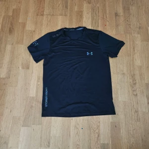Svart t-shirt från Under Armour - Svart t-shirt från Under Armour med korta ärmar och rund hals. Tillverkad i lätt och snabbtorkande polyester, perfekt för träning. Diskret logga på bröstet och tryck på ärmen samt längs sidan. Snygg och sportig stil som passar gymmet. Pris kan diskuteras!!