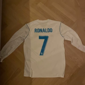 Real Madrid långärmad Ronaldo-tröja - Säljer en långärmad Real Madrid fotbollströja från Adidas med Ronaldo 7 på ryggen. Tröjan är vit med blå detaljer, klubbmärke på bröstet och Fly Emirates-tryck framtill. Klassisk V-ringning och tre ränder på axlarna. Ej inköpt från Real Madrid shopen. Fly emirates sponsorn börjar försvinna.