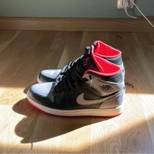 Nike Air Jordan 1 Mid Svart/Röd/Grå - Säljer ett par Nike Air Jordan 1 Mid i svart, grått och vitt med röda detaljer och röd yttersula. Skorna har klassisk snörning, ikonisk Jordan-logga på sidan och är tillverkade i skinn med textilfoder. Perfekta för dig som vill sticka ut med en clean och sportig look.