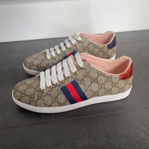Gucci ace - Snygga Gucci sneakers med klassiskt GG-monogram i beige och brunt. Skorna har vita snören, röd och blå rand på sidan samt metallicröd och metallicblå häl med Gucci-logga. Yttermaterial i canvas och detaljer i skinn. Perfekta för dig som vill sticka ut med lyxig streetstyle.