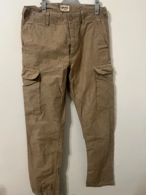 Beiga cargopants från Bad West - Säljer ett par beiga cargopants från Bad West med stora sidofickor och klassisk cargostil. Byxorna har normal passform och är tillverkade i bomull, perfekta för en avslappnad och cool look. Stängs med knapp och dragkedja.