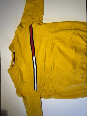 Gul tröja från Tommy Hilfiger M - Gul långärmad tröja från Tommy Hilfiger i storlek M. Tröjan har en rund halsringning och en randig detalj i rött, vitt och blått över bröstet samt i halsen. Tillverkad i mjuk bomull, perfekt för dig som gillar färg och klassisk stil.