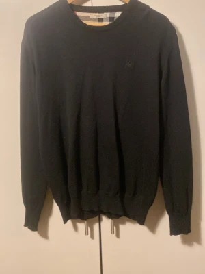 Svart stickad tröja från Burberry - Säljer en svart stickad tröja från Burberry med klassiskt rutigt mönster på axlar och armbågslappar. Tröjan har rund halsringning, ribbade muddar och en diskret Burberry-logga på bröstet. Perfekt för dig som gillar stilrena detaljer och premiumkänsla.