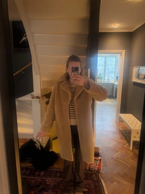 Beige teddyjacka från Stand Studio - Säljer en beige teddyjacka från Stand Studio med klassisk krage och knäppning framtill. Jackan är lång och har en oversized passform som ger en cool och avslappnad vibe. Perfekt för kyliga dagar när du vill vara både varm och snygg.